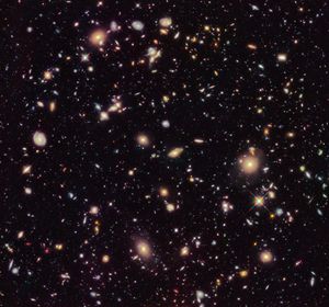 Image is filled with distant galaxies in colors of yellow-gold, red, blue, white. The whole image has a reddish tint.